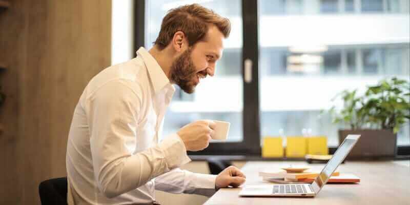 man-holding-teacup-infront-of-laptop-on-top-of-table-inside